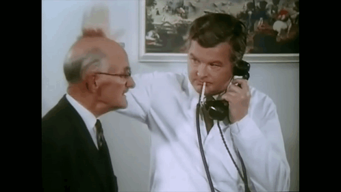 A Man In A White Coat Is Talking On A Phone With Another Man GIF