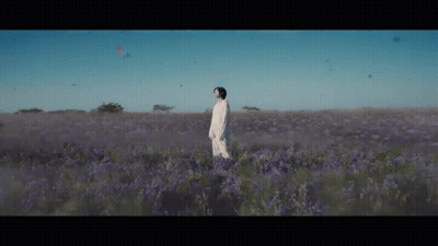 A Man In A White Dress Is Standing In A Field Of Purple Flowers . GIF