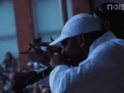 A Man In A White Hat Is Singing Into A Microphone In Front Of A Crowd And The Word Noise Is Visible In The Corner GIF