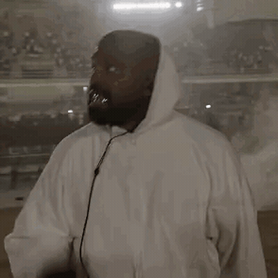 A Man In A White Hoodie Is Holding A Microphone In Front Of A Crowd . GIF