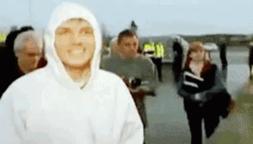 A Man In A White Hoodie Is Smiling In Front Of A Crowd Of People . GIF