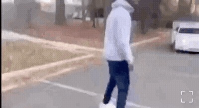A Man In A White Hoodie Is Walking Down A Street Next To A Car . GIF
