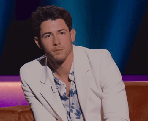 A Man In A White Jacket And Floral Shirt Is Sitting On A Brown Couch GIF