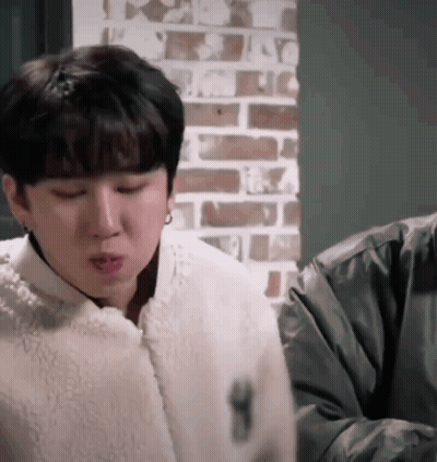 A Man In A White Jacket Is Sitting In Front Of A Brick Wall And Making A Funny Face . GIF