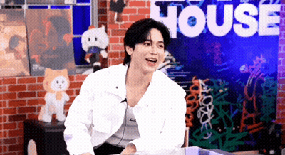 A Man In A White Jacket Is Sitting In Front Of A Brick Wall With The Word House On It . GIF