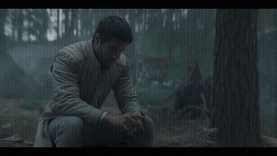 A Man In A White Jacket Sits In The Woods With His Hands On His Knees GIF