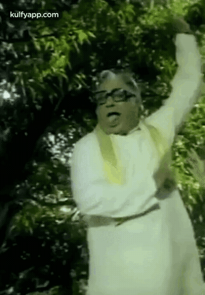 A Man In A White Robe And Glasses Is Standing In Front Of A Tree With His Arms In The Air . GIF