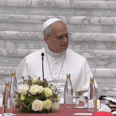 A Man In A White Robe Is Sitting At A Table With Bottles Of Water And Flowers GIF