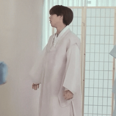 A Man In A White Robe Is Standing In Front Of A Room Divider Holding A Blue Shirt . GIF