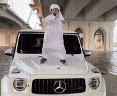 A Man In A White Robe Is Standing On The Hood Of A White Mercedes GIF