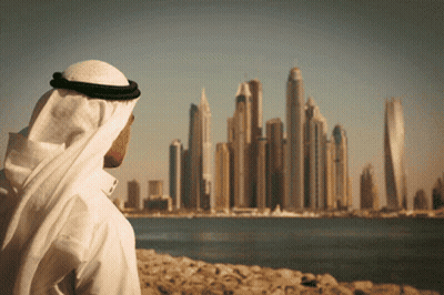 A Man In A White Robe Looks Out Over A Body Of Water At A City Skyline GIF