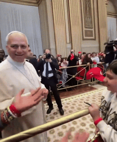 A Man In A White Robe Stands In Front Of A Crowd While A Boy In A Red Hat Stands Behind Him GIF