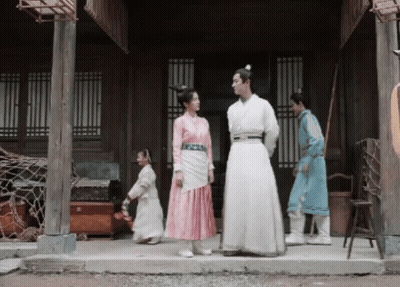 A Man In A White Robe Stands Next To A Woman In A Pink Dress On A Porch GIF