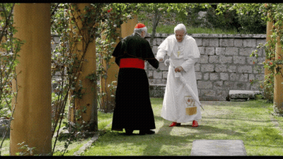 A Man In A White Robe With A Cross On It Stands Next To Another Man In A Black Robe GIF