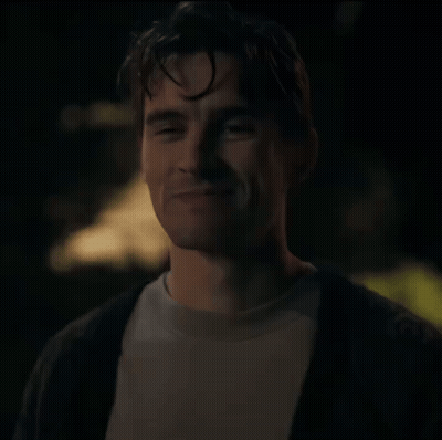 A Man In A White Shirt And A Black Cardigan Smiles At The Camera GIF