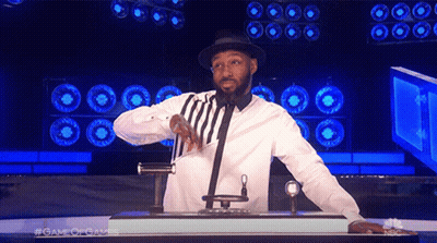 A Man In A White Shirt And Black Hat Stands In Front Of A Microphone With The Words Game Of Games Behind Him GIF