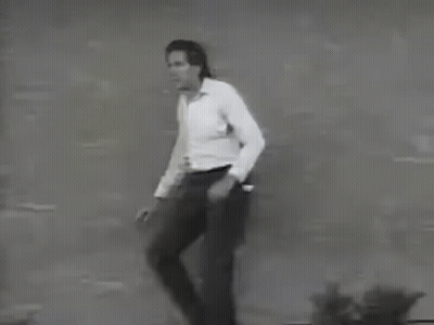 A Man In A White Shirt And Black Pants Is Standing In Front Of A Brick Wall . GIF