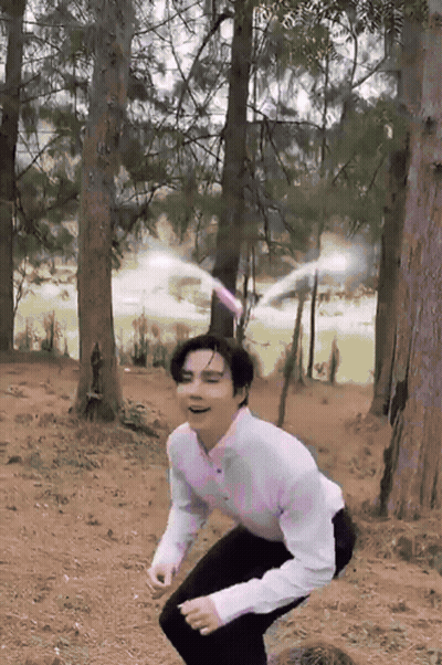A Man In A White Shirt And Black Pants Squats In The Woods GIF