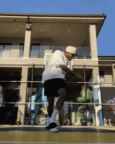 A Man In A White Shirt And Black Shorts Stands On A Basketball Court In Front Of A House GIF