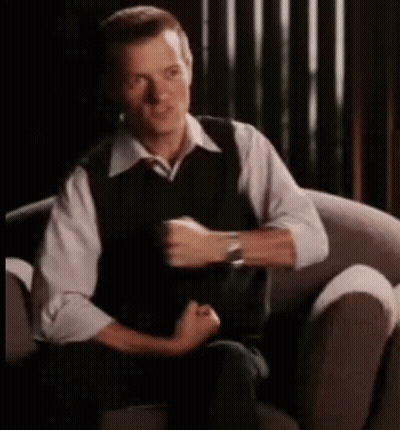 A Man In A White Shirt And Black Vest Is Sitting In A Chair . GIF