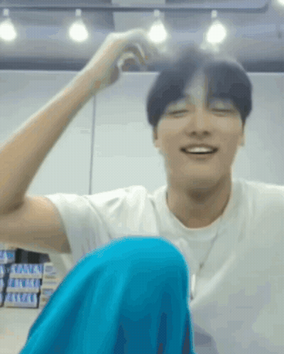 A Man In A White Shirt And Blue Pants Is Sitting On The Floor With His Legs Crossed And Smiling . GIF