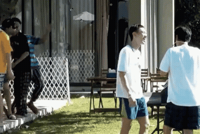 A Man In A White Shirt And Blue Shorts Is Standing In A Yard With Other People GIF