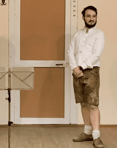 A Man In A White Shirt And Brown Shorts Stands In Front Of A Music Stand GIF