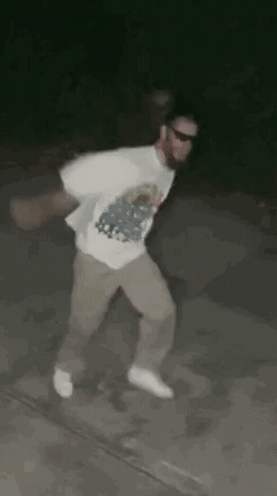 A Man In A White Shirt And Khaki Pants Is Dancing On A Sidewalk At Night . GIF