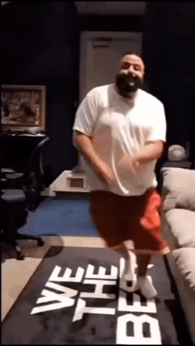 A Man In A White Shirt And Red Shorts Is Dancing On A Mat That Says We The Best . GIF