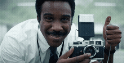 A Man In A White Shirt And Tie Is Holding A Camera And Giving A Thumbs Up GIF