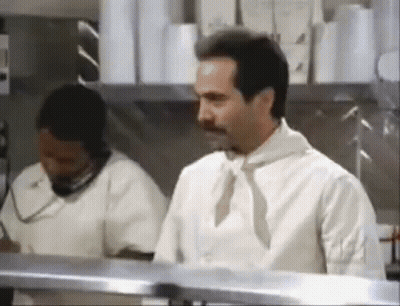 A Man In A White Shirt And Tie Is Standing In A Kitchen Next To A Woman Talking On A Phone . GIF