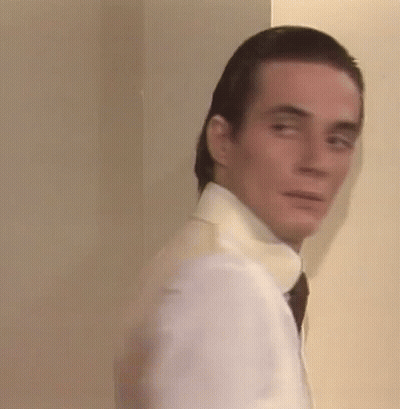 A Man In A White Shirt And Tie Is Standing Next To A Wall And Looking At The Camera . GIF