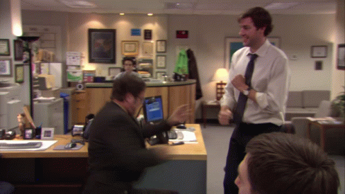 A Man In A White Shirt And Tie Stands In Front Of A Man Sitting At A Desk GIF