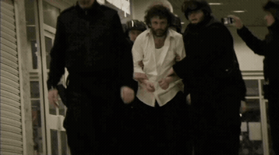 A Man In A White Shirt Is Being Led By A Group Of Police Officers GIF