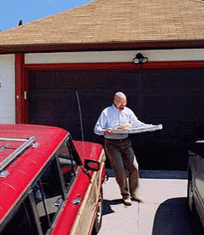 A Man In A White Shirt Is Carrying A Pizza In Front Of A Garage Door GIF