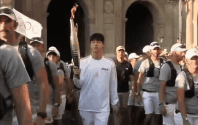 A Man In A White Shirt Is Carrying A Torch In Front Of A Crowd Of People . GIF