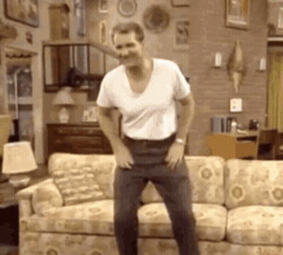 A Man In A White Shirt Is Dancing In Front Of A Couch . GIF