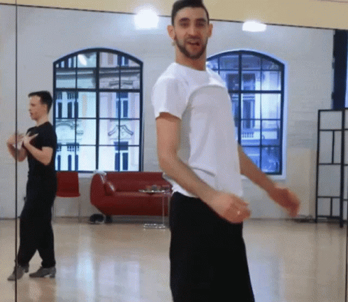 A Man In A White Shirt Is Dancing In Front Of A Large Mirror GIF