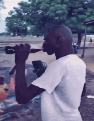 A Man In A White Shirt Is Drinking From A Bottle Outside GIF