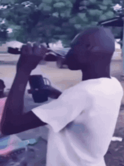 A Man In A White Shirt Is Drinking From A Bottle Outside GIF