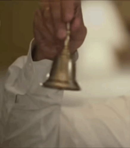 A Man In A White Shirt Is Holding A Gold Bell In His Hand . GIF