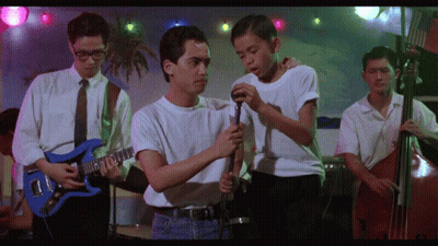 A Man In A White Shirt Is Holding A Microphone While Another Man Plays A Guitar GIF