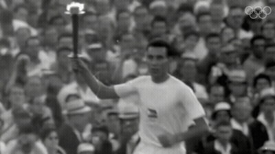 A Man In A White Shirt Is Holding A Torch In Front Of A Crowd With The Olympic Rings In The Background GIF