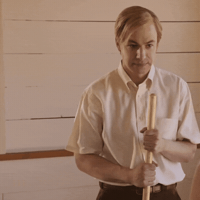 A Man In A White Shirt Is Holding A Wooden Stick In His Right Hand GIF