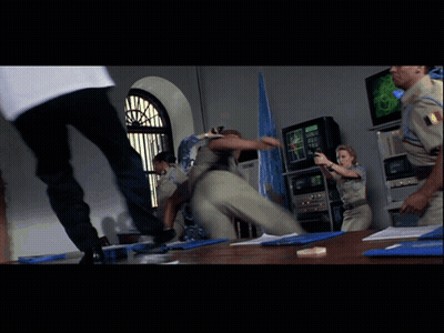 A Man In A White Shirt Is Kicking Another Man In A Room With Other People GIF