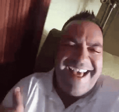 A Man In A White Shirt Is Laughing With His Mouth Open While Sitting In A Chair . GIF