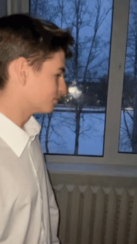 A Man In A White Shirt Is Looking Out A Window GIF
