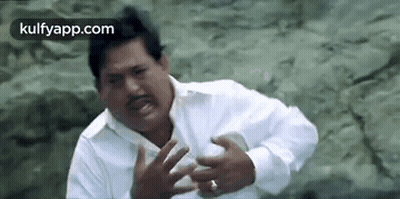 A Man In A White Shirt Is Making A Funny Face While Sitting On A Rock . GIF