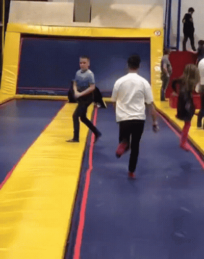 A Man In A White Shirt Is Running On A Trampoline While A Boy In A Blue Shirt Watches GIF