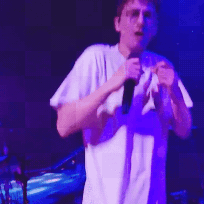 A Man In A White Shirt Is Singing Into A Microphone On Stage GIF